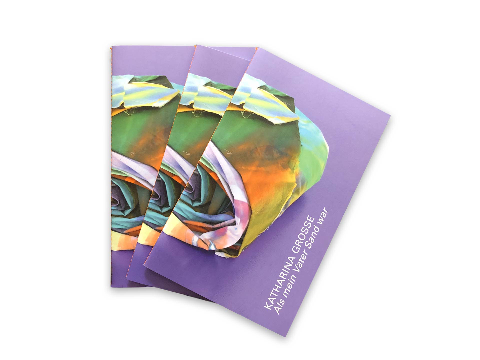 Foto von drei Booklets. Diese liegen gefächert und zeigen ein Motiv mit bunten Stoffen die zu eine Rolle zusammengerollt sind.